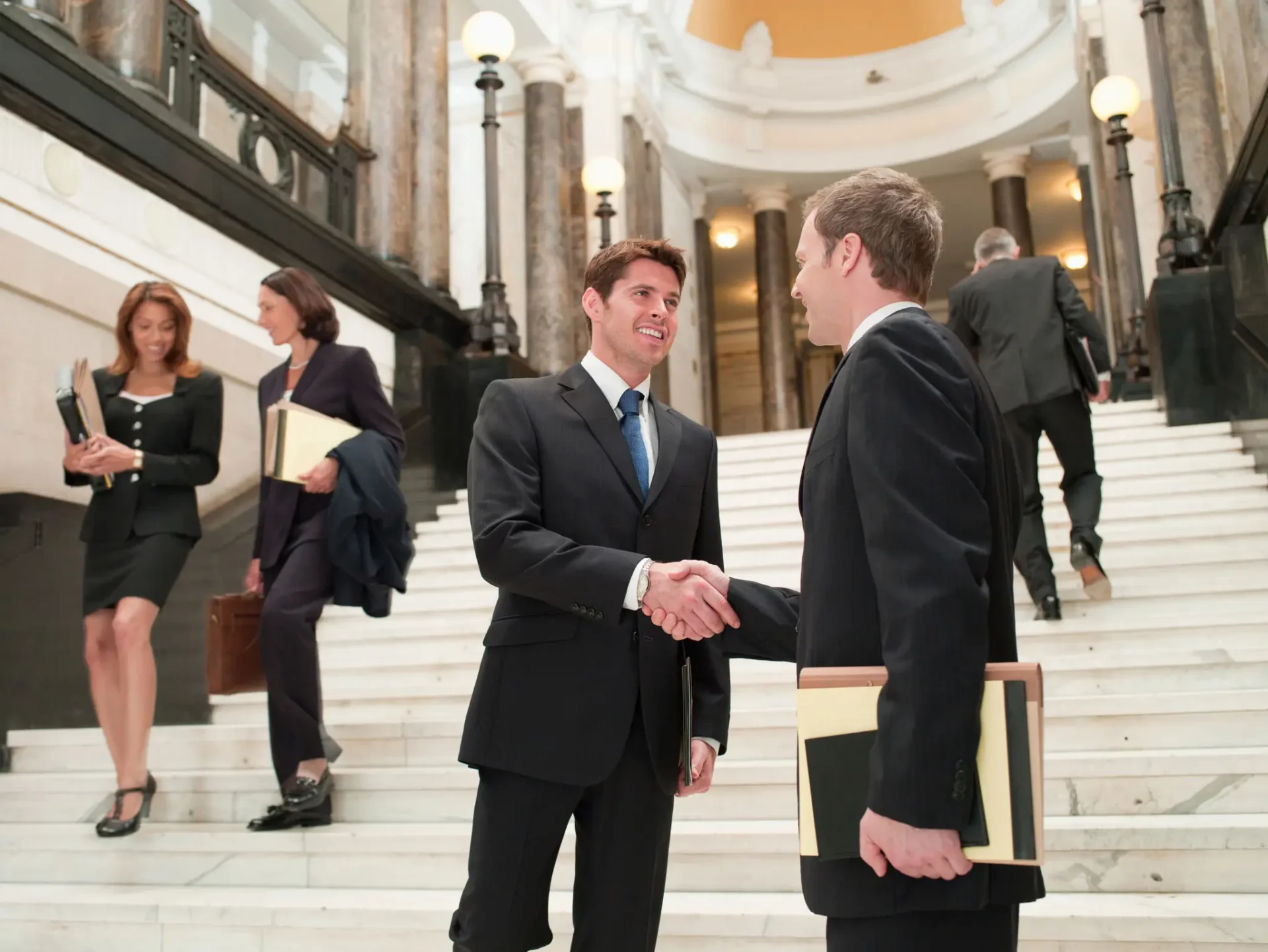 Two businessmen shaking hands on a grand staircase in a formal setting.